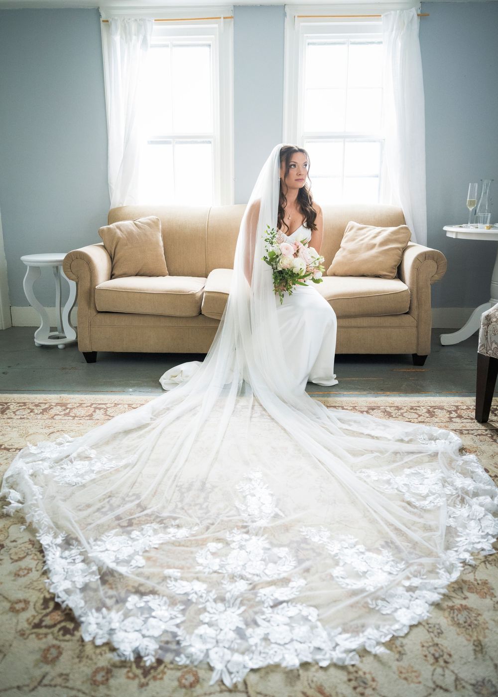 Bride in stunning lace cathedral veil and white gown sits gracefully on beige sofa holding bridal bouquet.