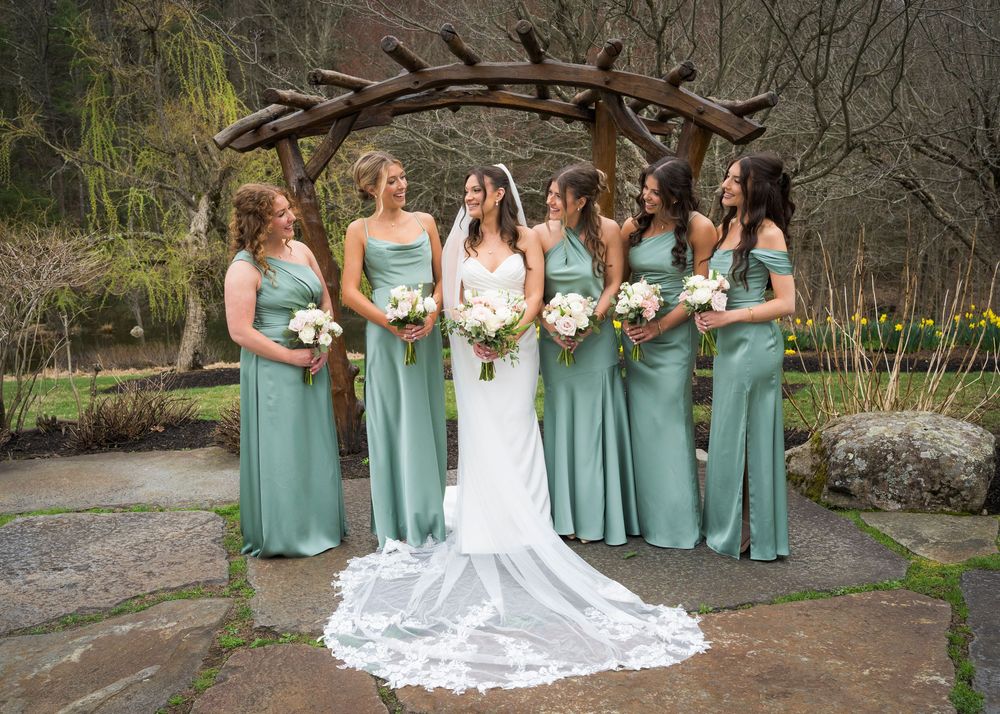 Bride with six bridesmaids in sage green gowns holding white bouquets under rustic wooden arch.