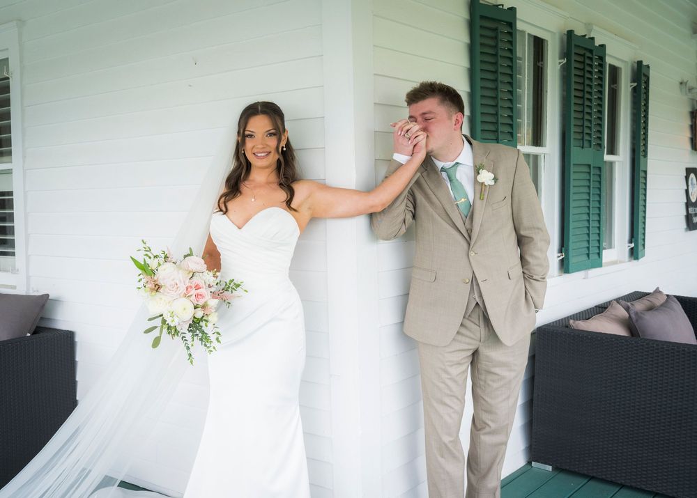 Bride in white gown and groom in tan suit share emotional first look moment on white farmhouse porch.