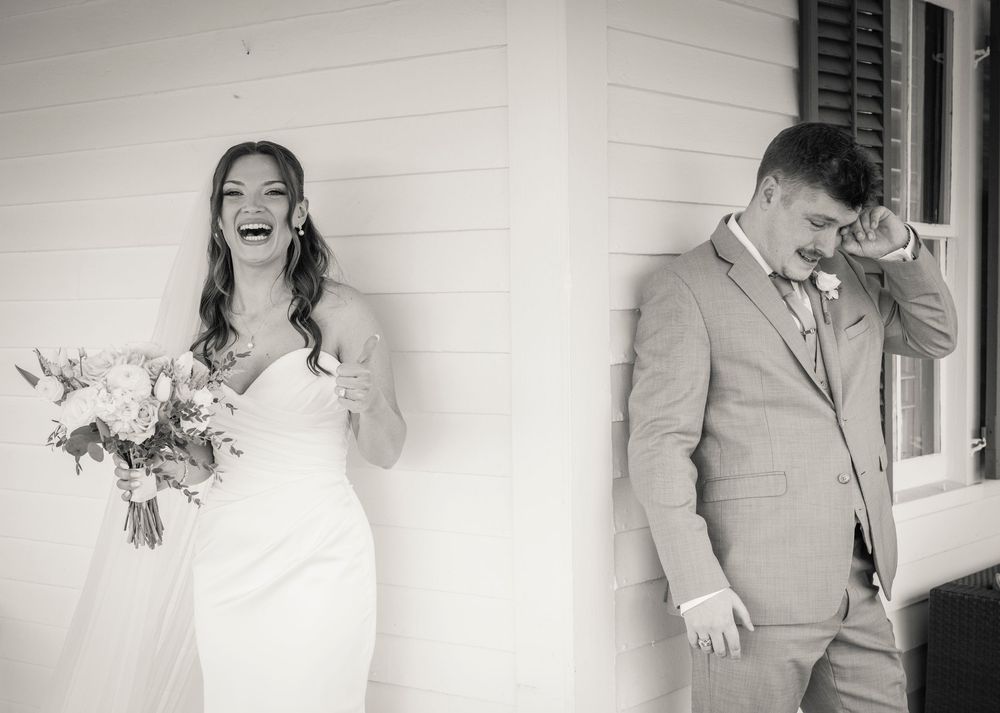 Bride laughs joyfully while groom wipes tears during an emotional first look at their outdoor wedding.