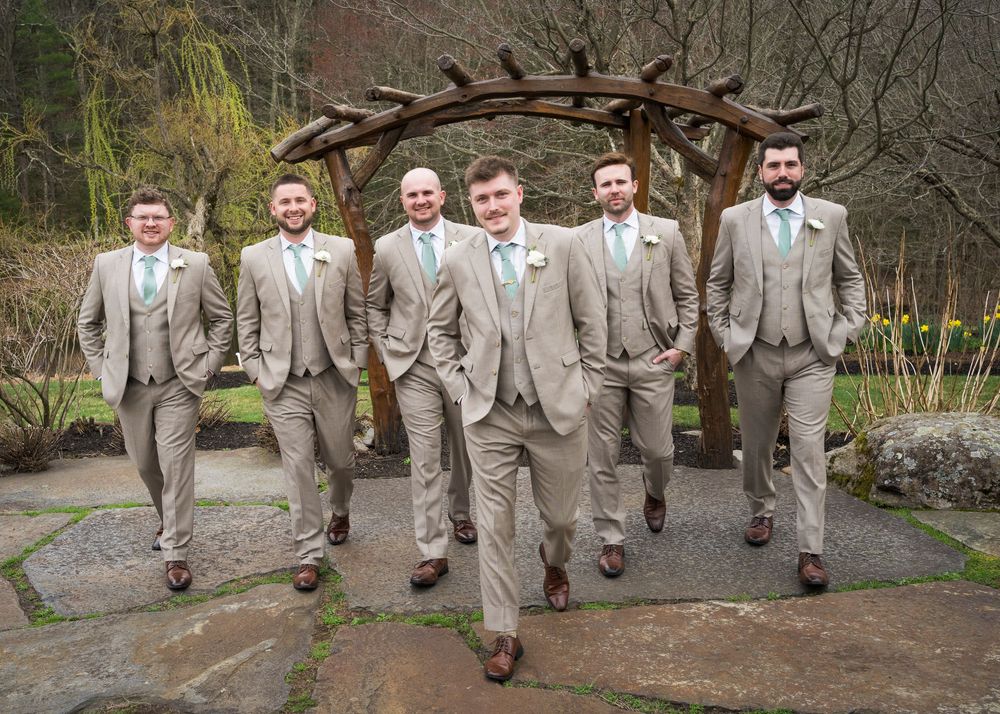 Groom and five groomsmen in tan suits walking confidently under rustic wooden arch outdoors.