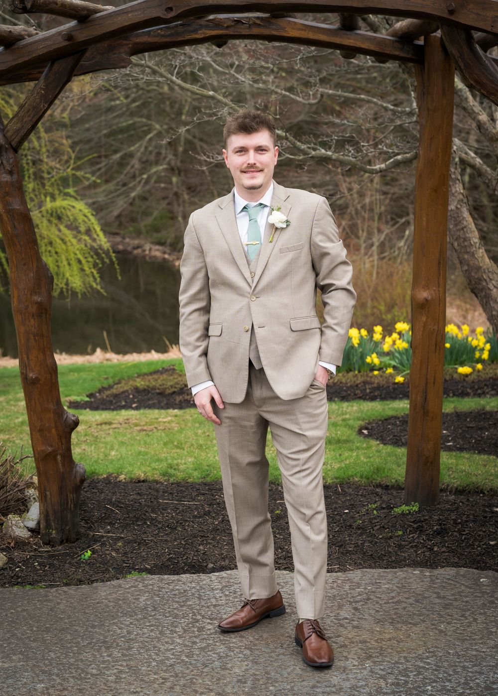Groom in tan suit with boutonniere poses under rustic wooden arbor with daffodils at Harrington Farm.