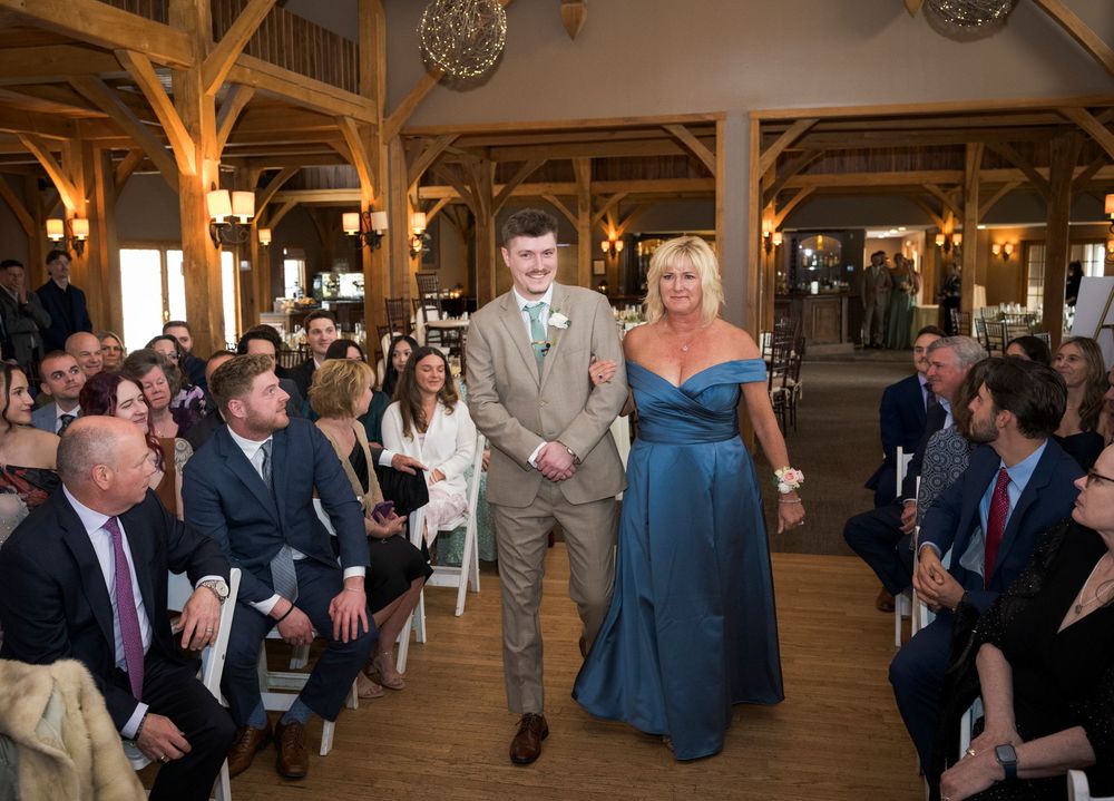 Groom in tan suit walks down wedding aisle with mother in blue gown at rustic barn venue.