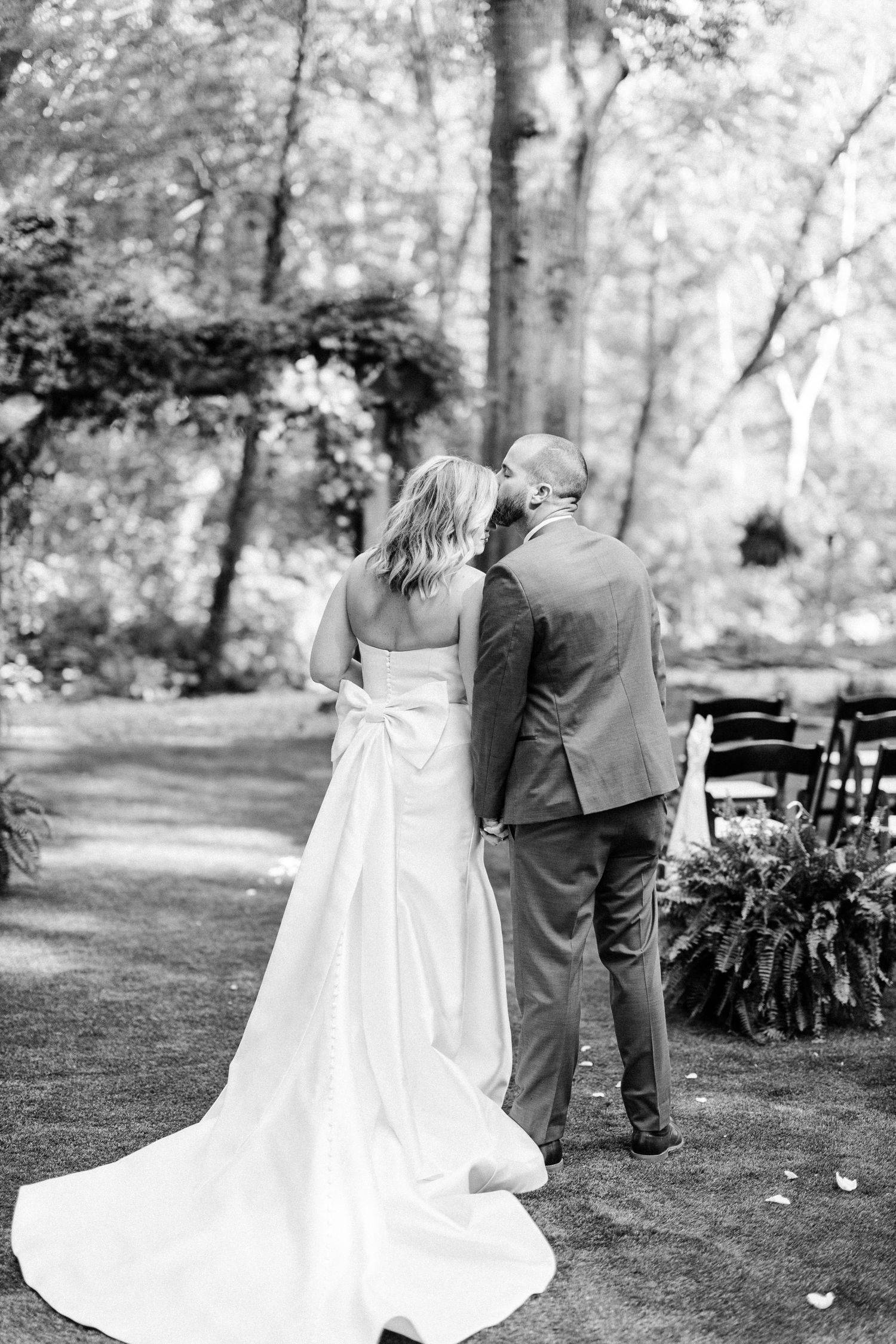 A black and white wedding photo showing a couple from behind standing in a garden setting with chairs and greenery.