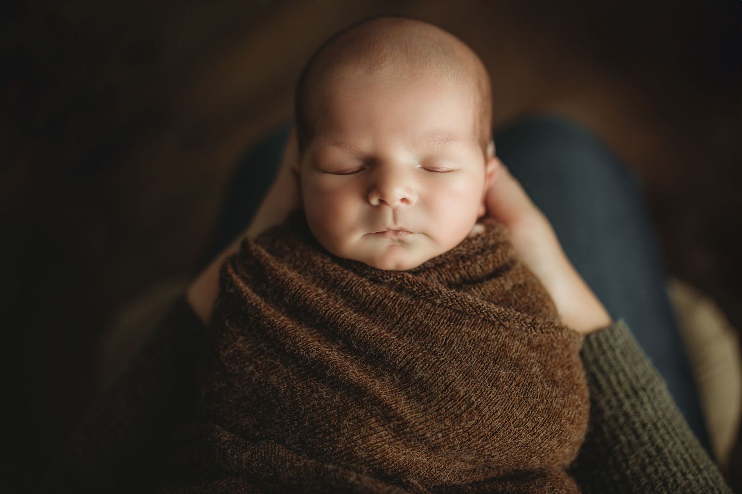 Cullman, Alabama newborn photographer. Sleeping newborn baby wrapped in soft brown blanket rests peacefully in dark studio lighting.