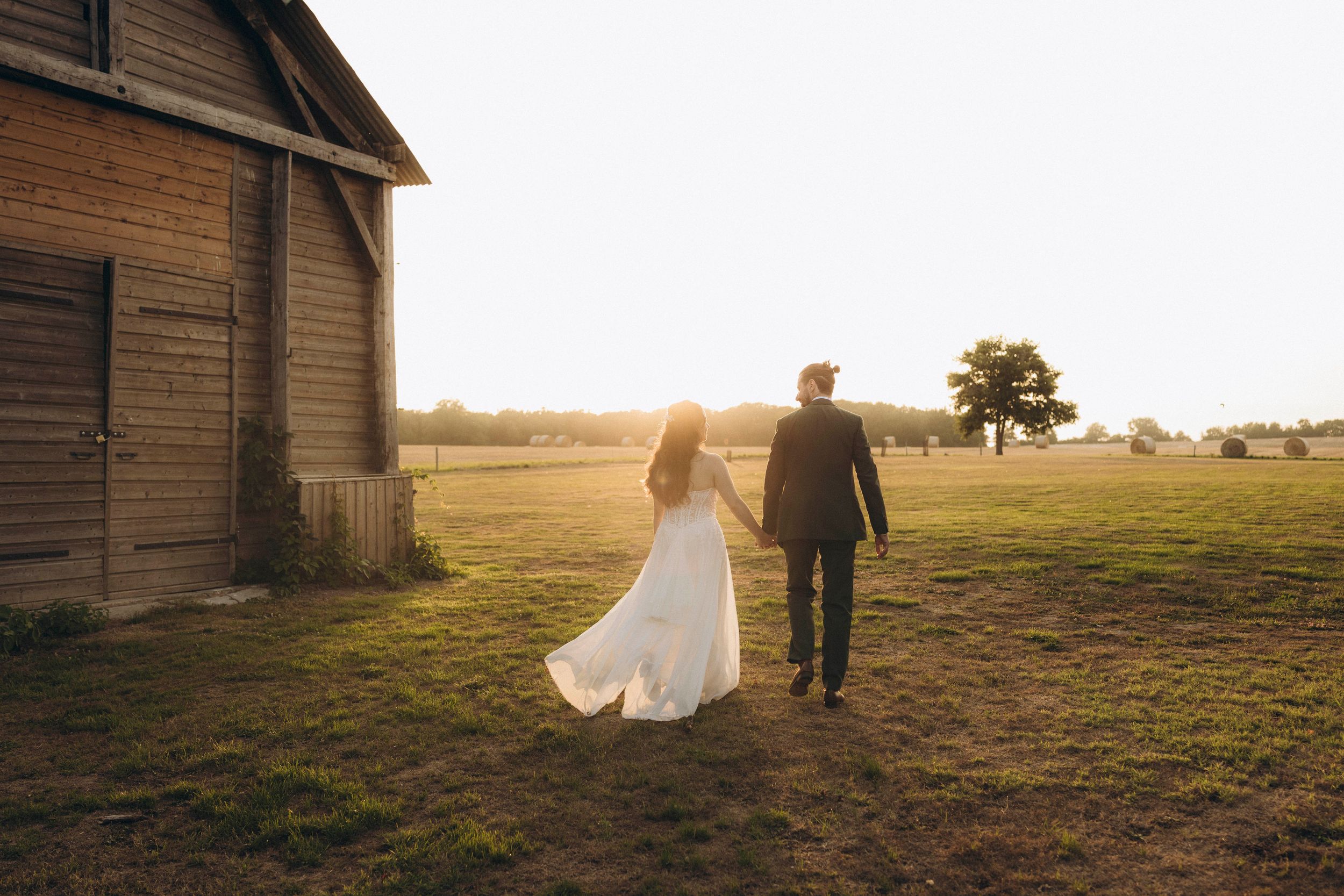 A couple walks hand in hand through a golden field at sunset near a rustic wooden barn.