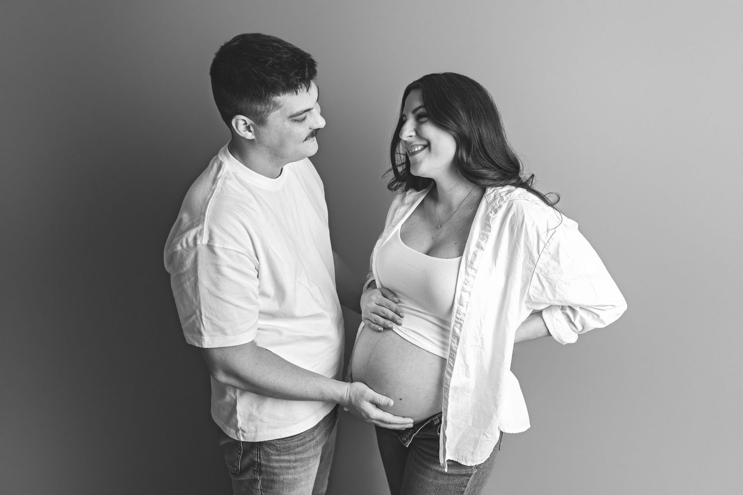 Expectant couple in casual attire standing barefoot against white wall during maternity photoshoot.