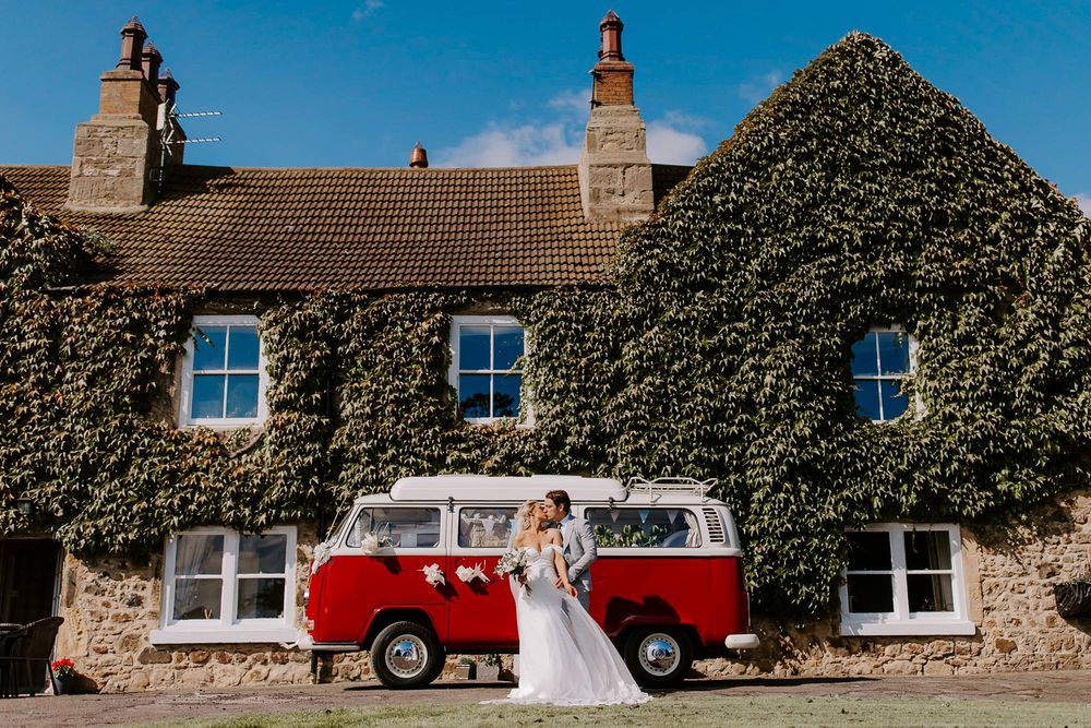 A vintage red and white VW bus parked in front of an ivy-covered stone cottage on a sunny day.