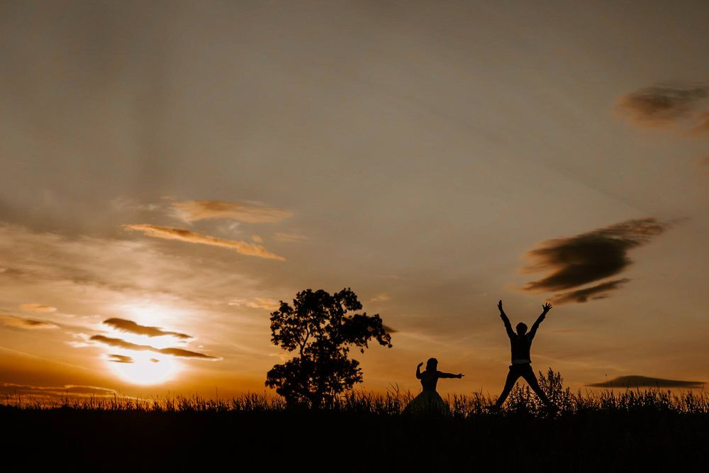Silhouettes of people jumping against dramatic sunset sky with trees in background.