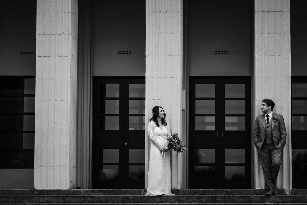 A black and white wedding photo of a couple standing between elegant columns in front of dark wooden doors.