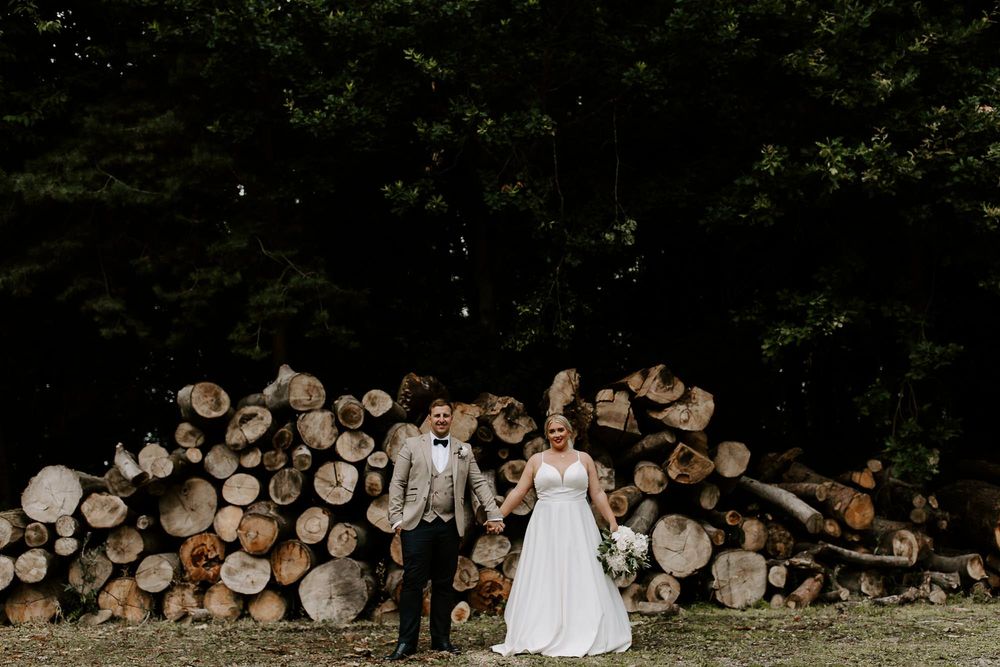 A couple in wedding attire stands in front of stacked firewood logs in a rustic outdoor setting.