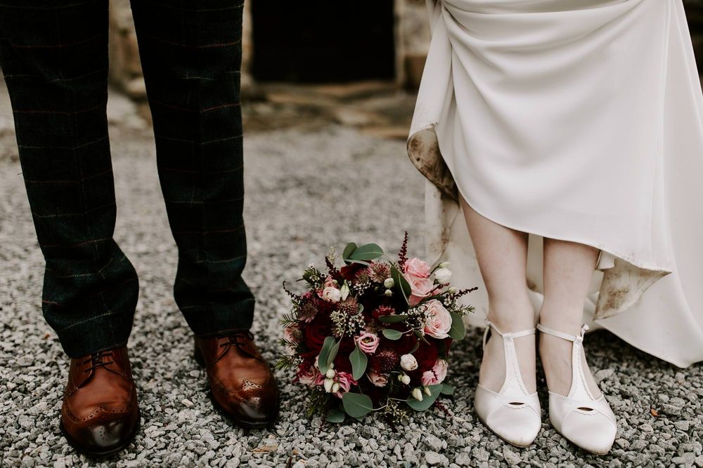 A wedding detail shot showing brown leather dress shoes and white bridal heels with a burgundy floral bouquet between them.