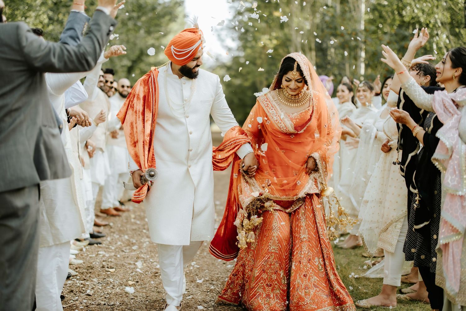 A festive wedding procession with guests in white clothing celebrating on a tree-lined path.