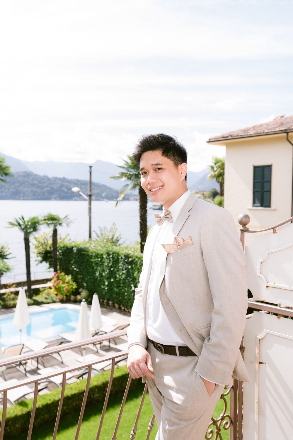 A person in a light gray suit stands on a terrace overlooking a lake and mountains with palm trees in the background.