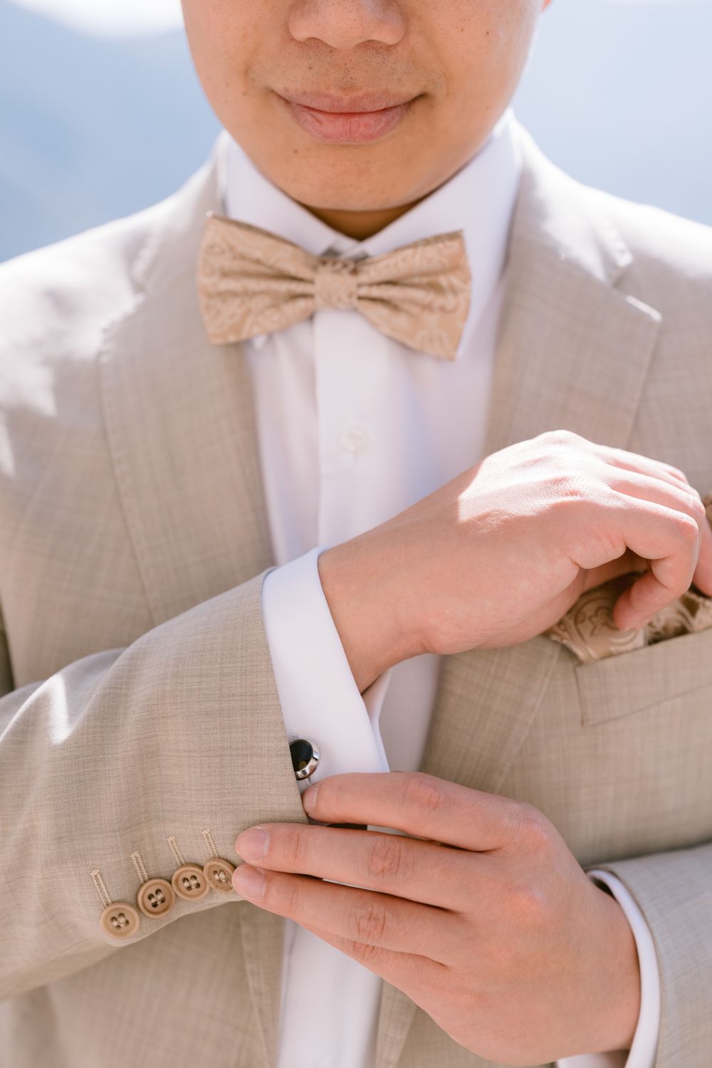 Close-up of hands adjusting a light pink bow tie against a white dress shirt and beige suit jacket.