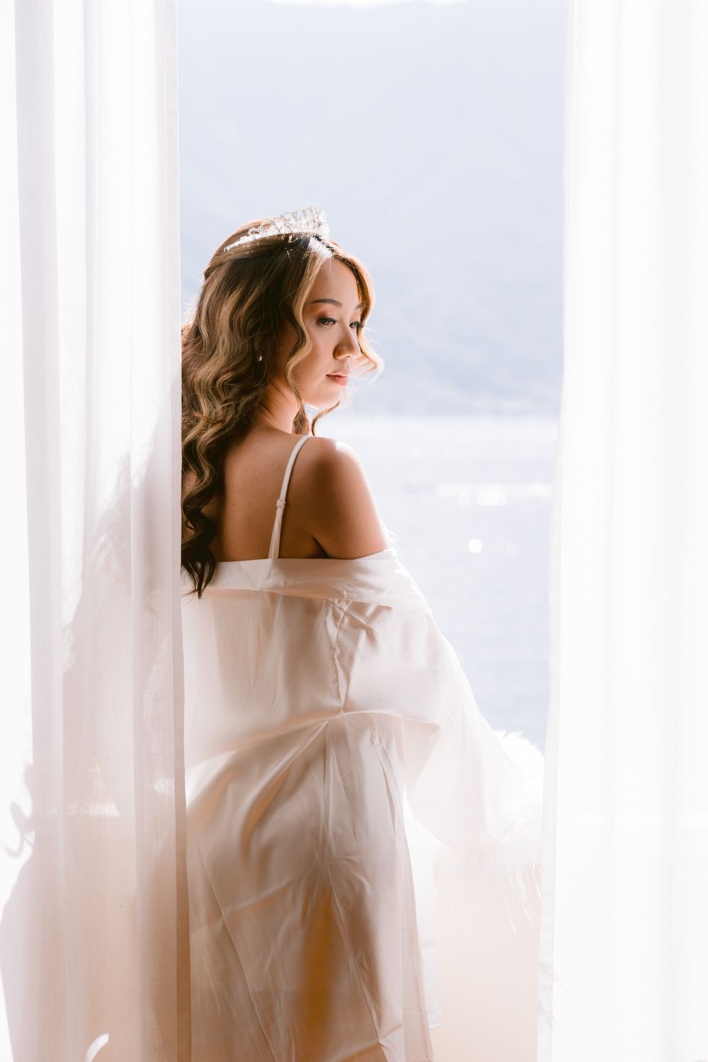 Romantic portrait by a window with soft lighting and flowing white fabric creating a dreamy atmosphere.