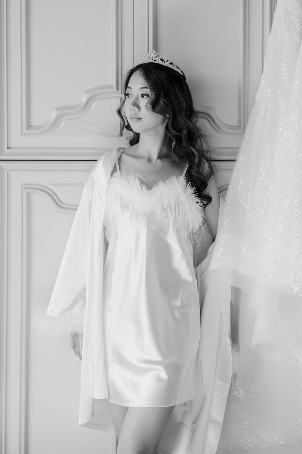 Black and white boudoir photo showing a figure in a delicate white silk slip dress and tiara against an ornate wall.