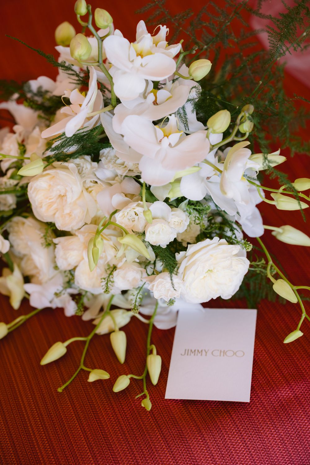 Elegant white floral arrangement with roses and orchids next to a Jimmy Choo card on a red textured surface.