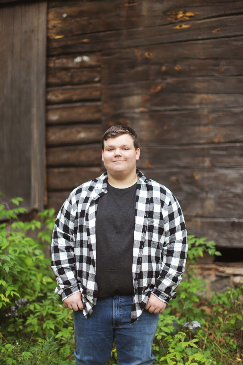 Someone wearing a black and white plaid flannel shirt and jeans poses in front of a rustic wooden wall with greenery.
