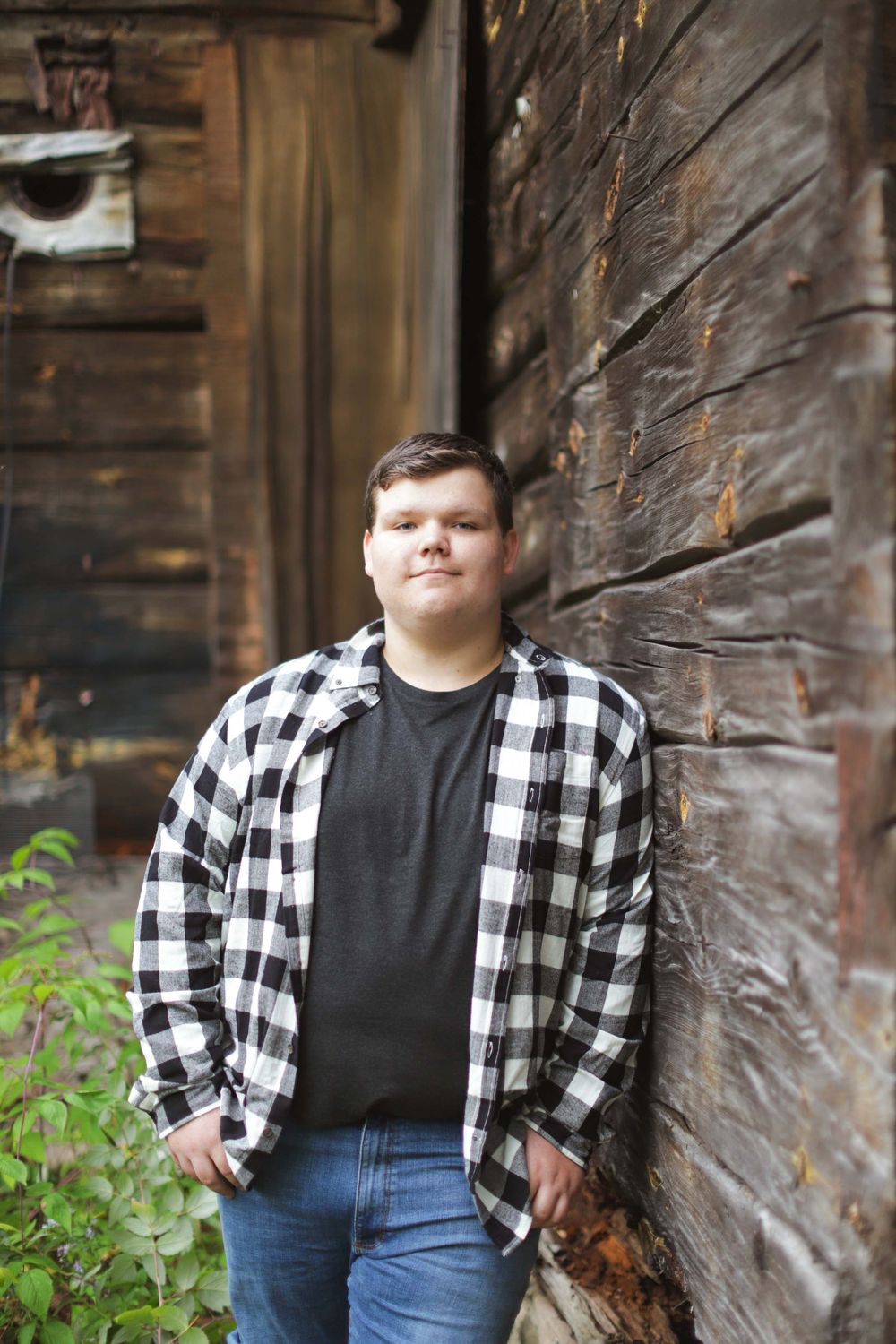 Person in black shirt and plaid flannel posing against rustic wooden wall in outdoor portrait setting.