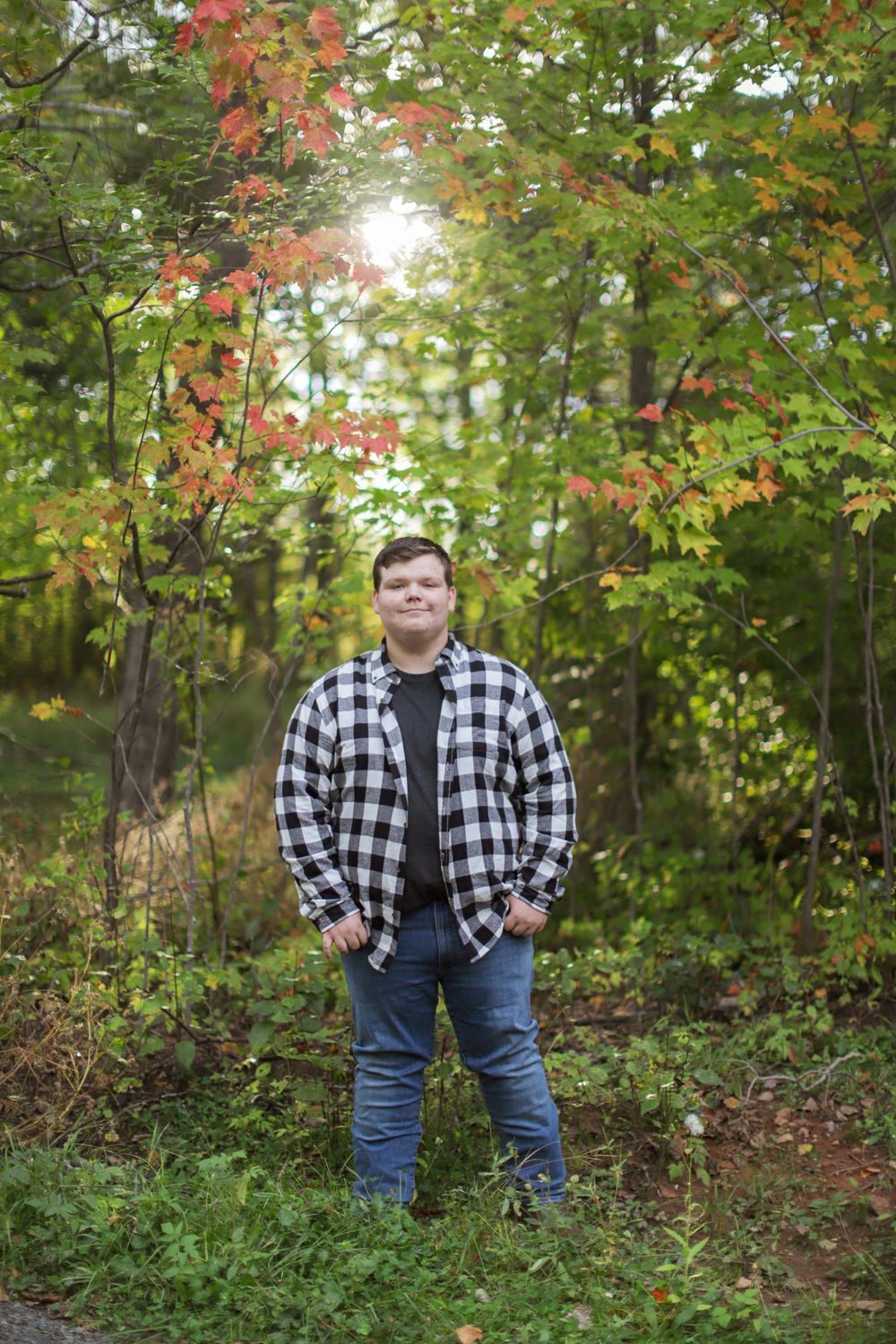 Person wearing black and white plaid shirt and jeans stands in autumn woodland setting with colorful foliage.