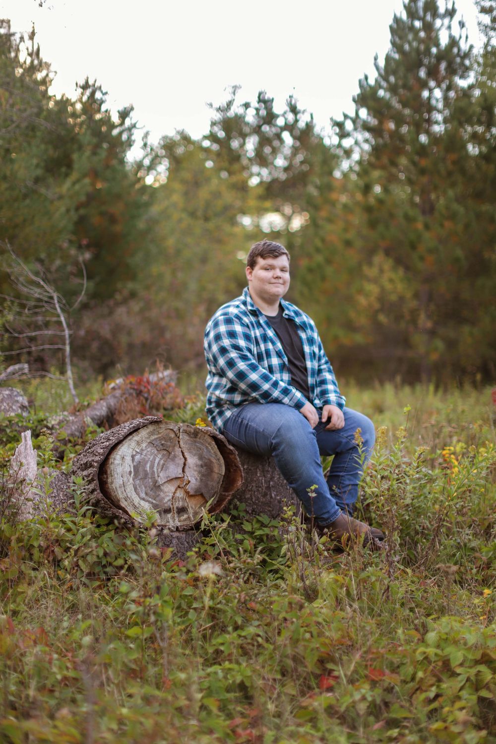 A rustic outdoor portrait scene with someone in a blue plaid shirt sitting on a fallen log among autumn foliage.