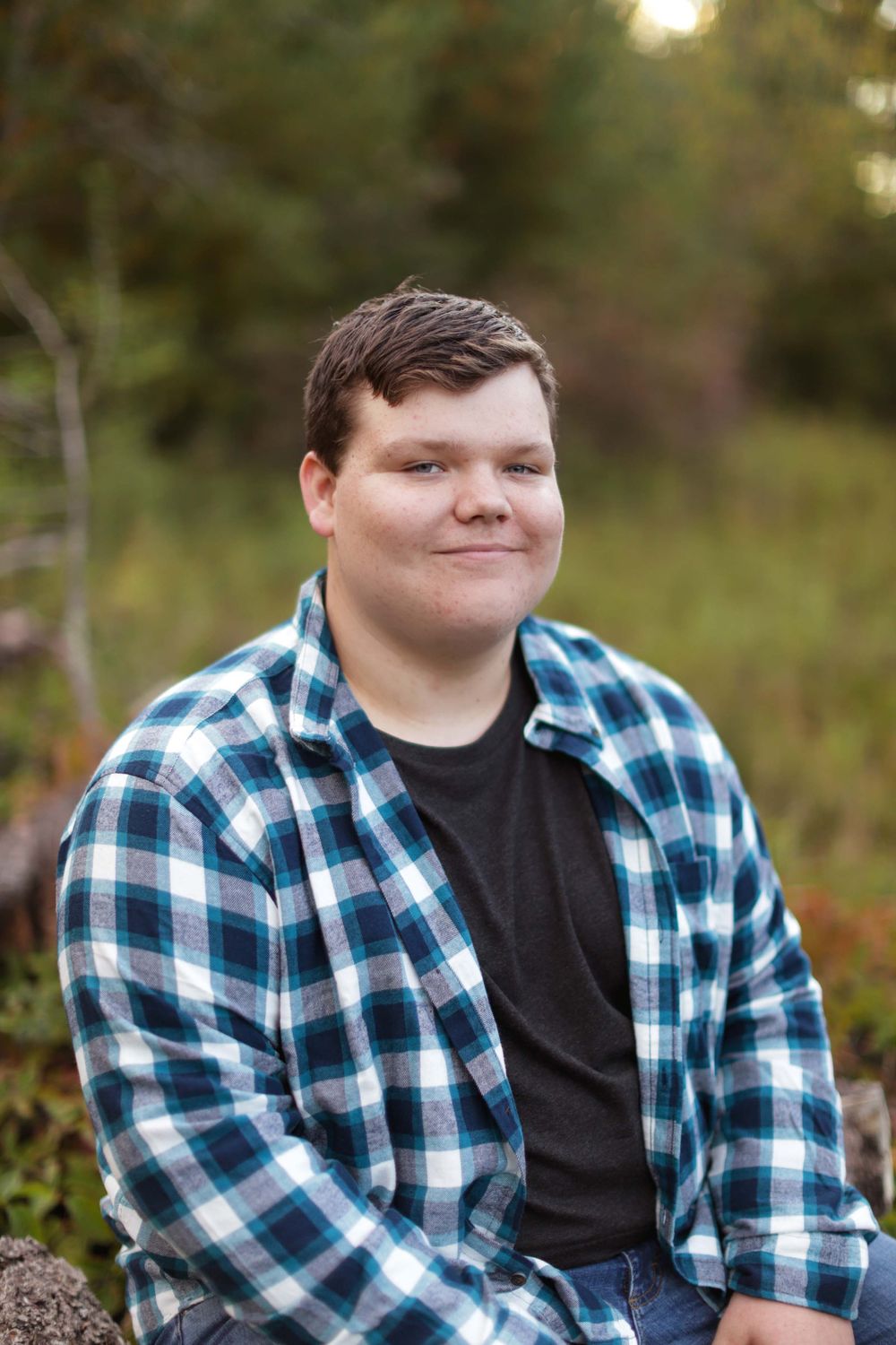 Portrait in natural setting with person wearing blue plaid shirt and black top against blurred outdoor background.