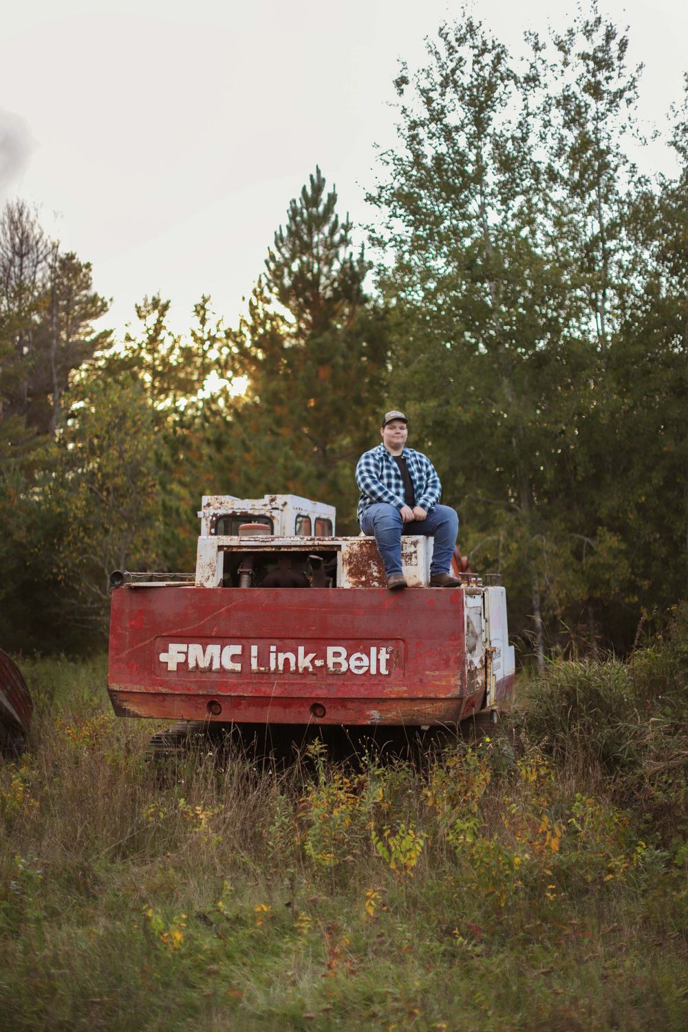 A rustic red FMC Link-Belt truck sits in a grassy field at sunset with pine trees in the background.