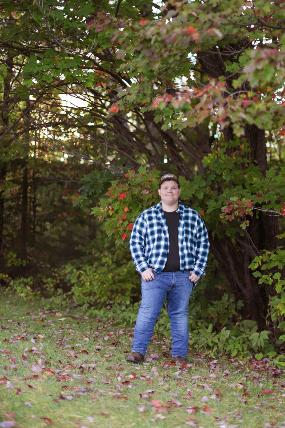 A casual outdoor scene with someone wearing denim and a blue plaid shirt standing among fall leaves and greenery.