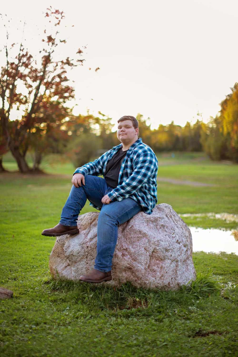 Person in plaid shirt and jeans relaxing on a large rock by a pond during golden hour.