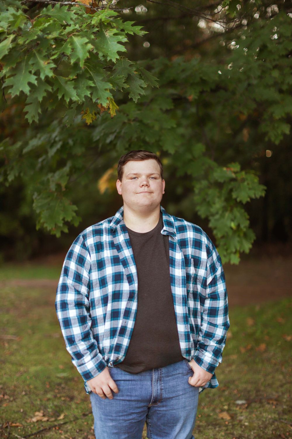 Person in blue plaid button-up and jeans poses confidently in front of fall trees and greenery.