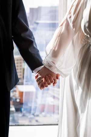 Close-up view of a couple holding hands against a bright window, with the groom in dark suit and bride in white dress.