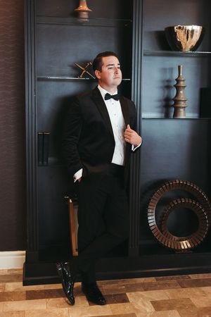 Man in formal black tuxedo and bow tie poses against dark wall with circular mirror in elegant interior setting.
