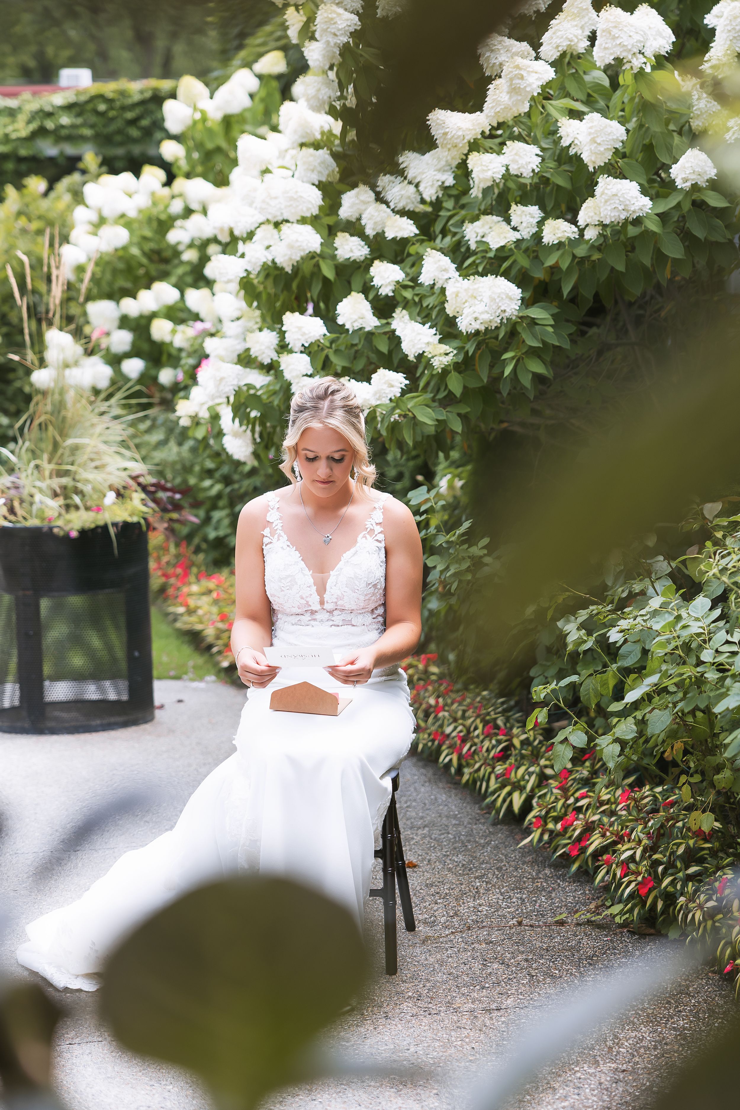 A serene garden scene with white roses framing a gravel path where someone in a white dress sits on a bench.