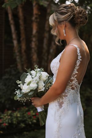 Side view of bride in elegant lace wedding dress holding white rose bouquet during outdoor garden ceremony.