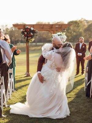 A romantic sunset wedding ceremony with a couple dipping under a wooden arch surrounded by white chairs on a grassy hill.