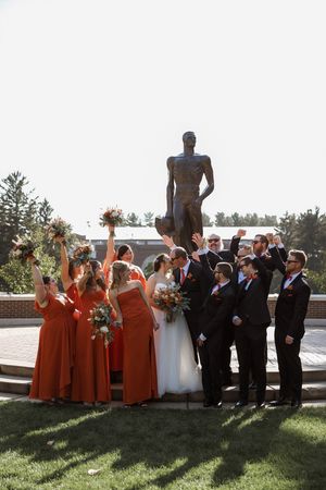 A wedding party in orange bridesmaid dresses and black suits poses for photos in front of a large statue on a sunny day.