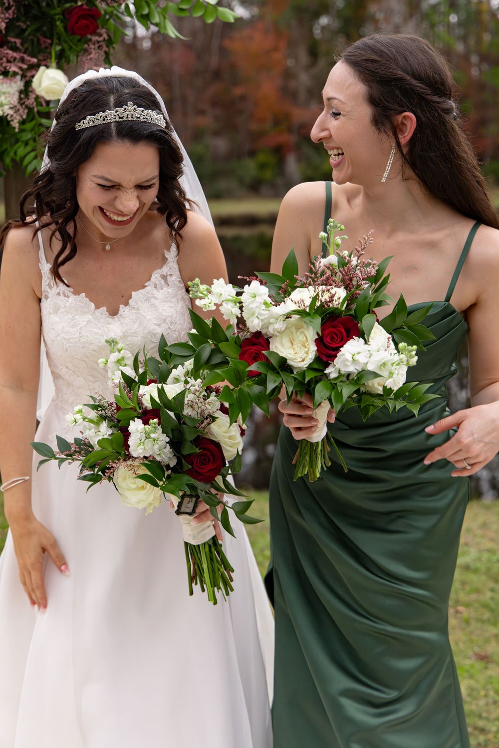 Wedding party members in emerald green dresses and white gown hold bouquets of red and white flowers in outdoor setting.