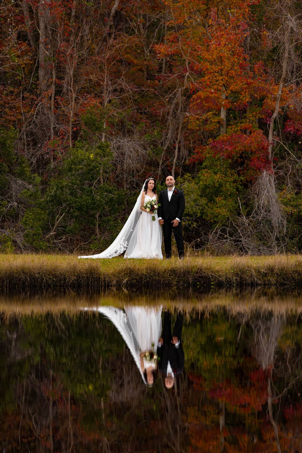 A wedding couple stands by a lake with stunning fall foliage creating a perfect reflection in the water.