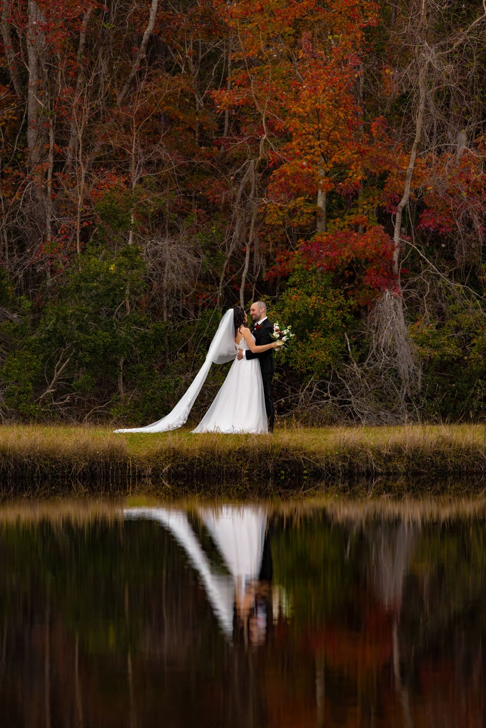 A couple in wedding attire stands by a tranquil lake surrounded by vibrant autumn foliage creating a stunning reflection.