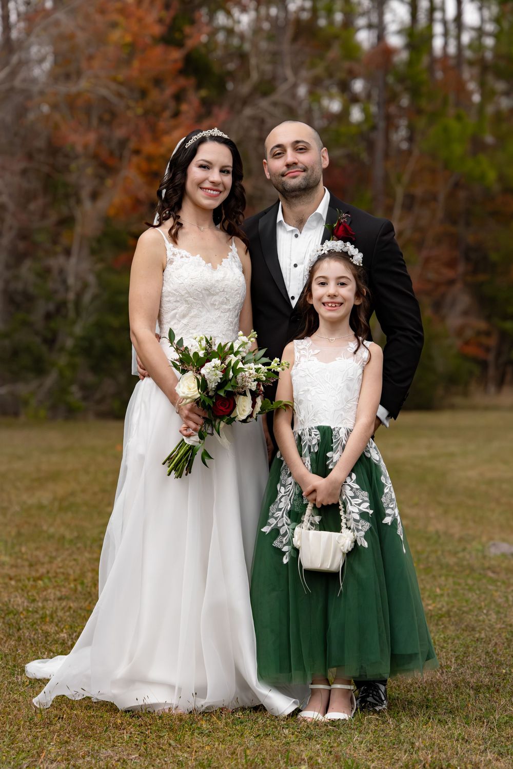 Wedding party poses outdoors in autumn with bride in white dress and flower girl in emerald green dress.