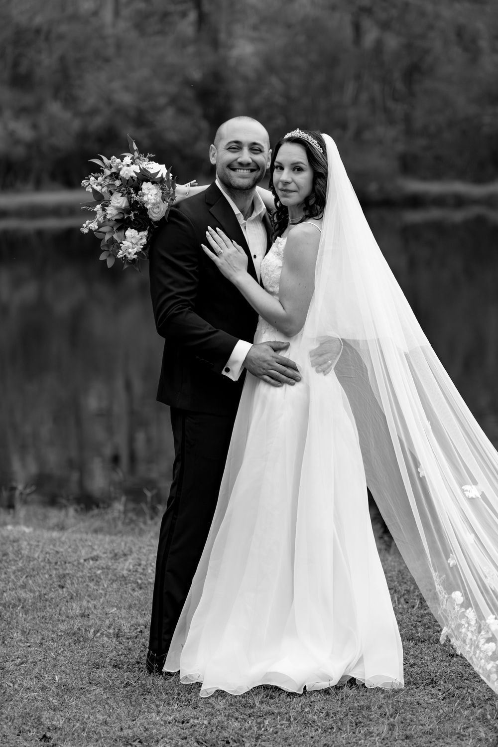 Black and white wedding portraits by a lake with flowing veil and elegant poses.