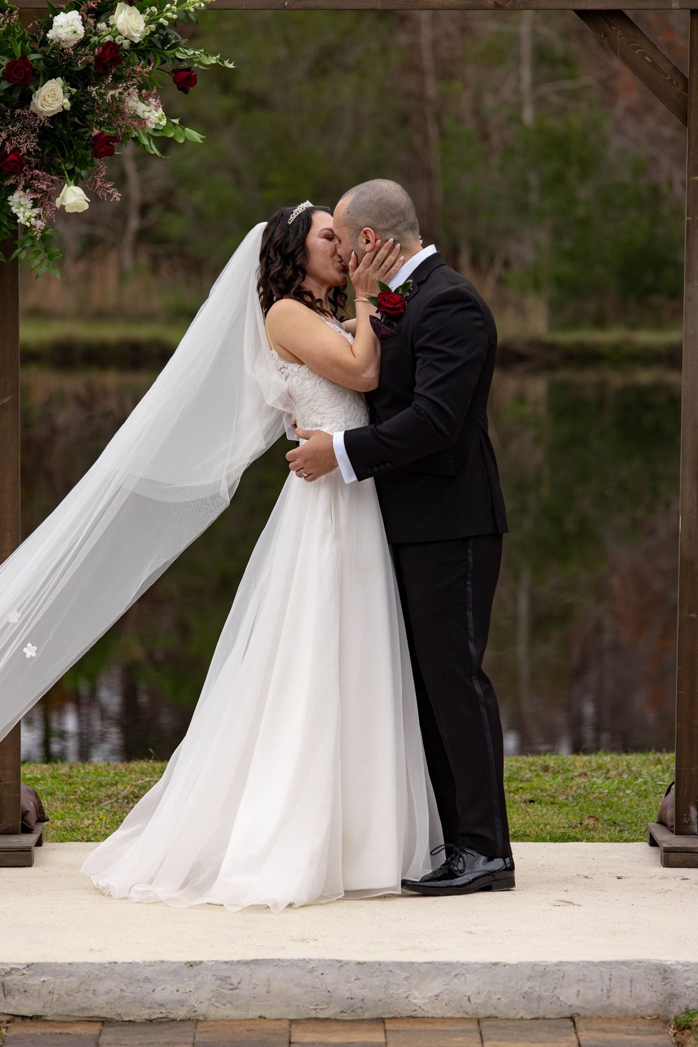 A romantic wedding kiss under a floral arch with a flowing white veil and elegant formal attire.