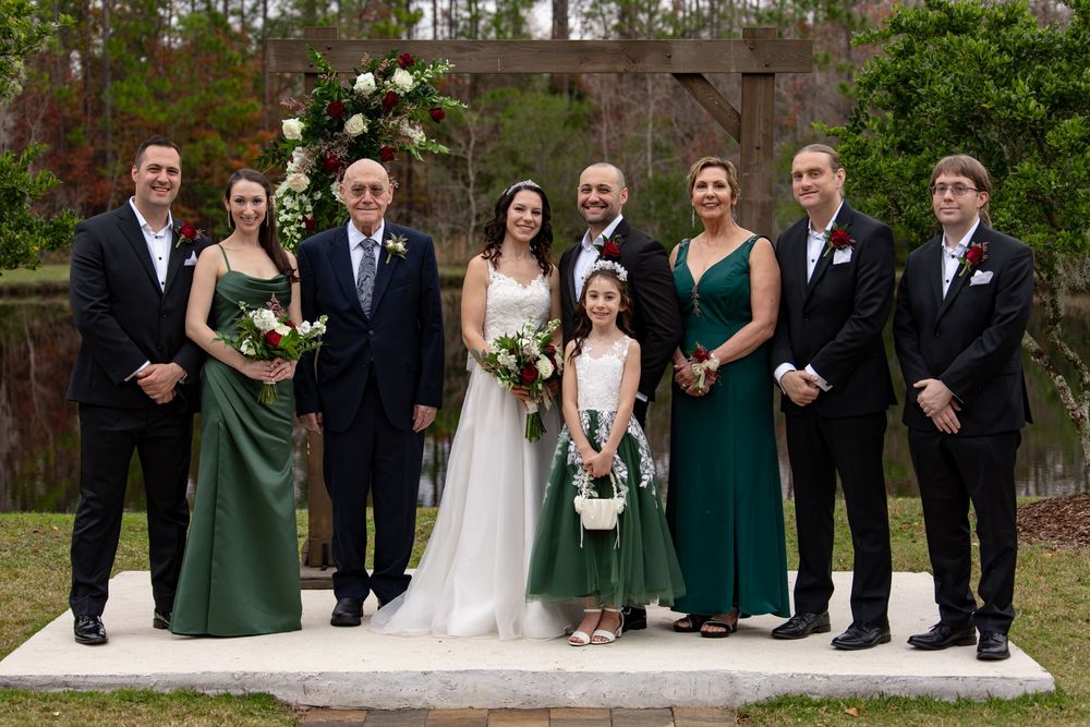 Wedding party in formal attire poses outdoors with bridesmaids wearing emerald green dresses and holding bouquets.