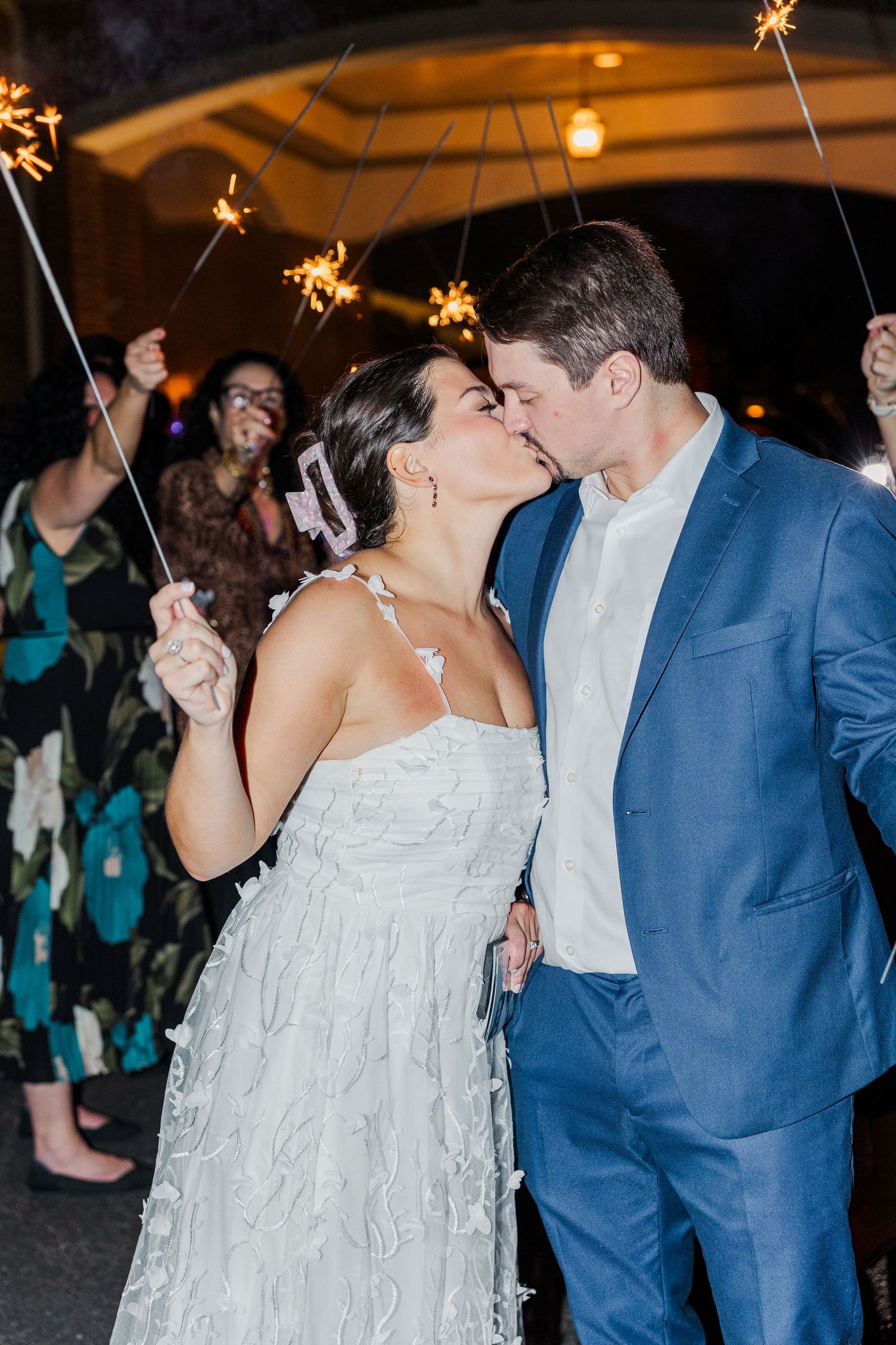 A romantic wedding moment captured as a couple shares a kiss while guests hold sparklers during an evening celebration.