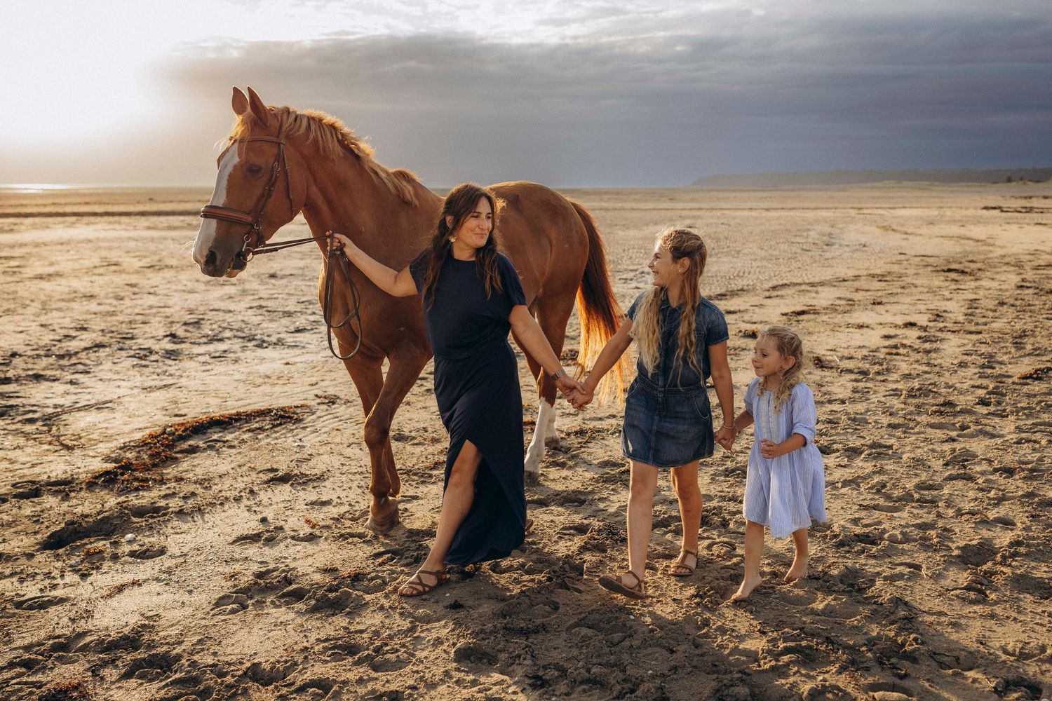 People and horses stand together on a sandy beach at sunset creating silhouettes against the dramatic sky.