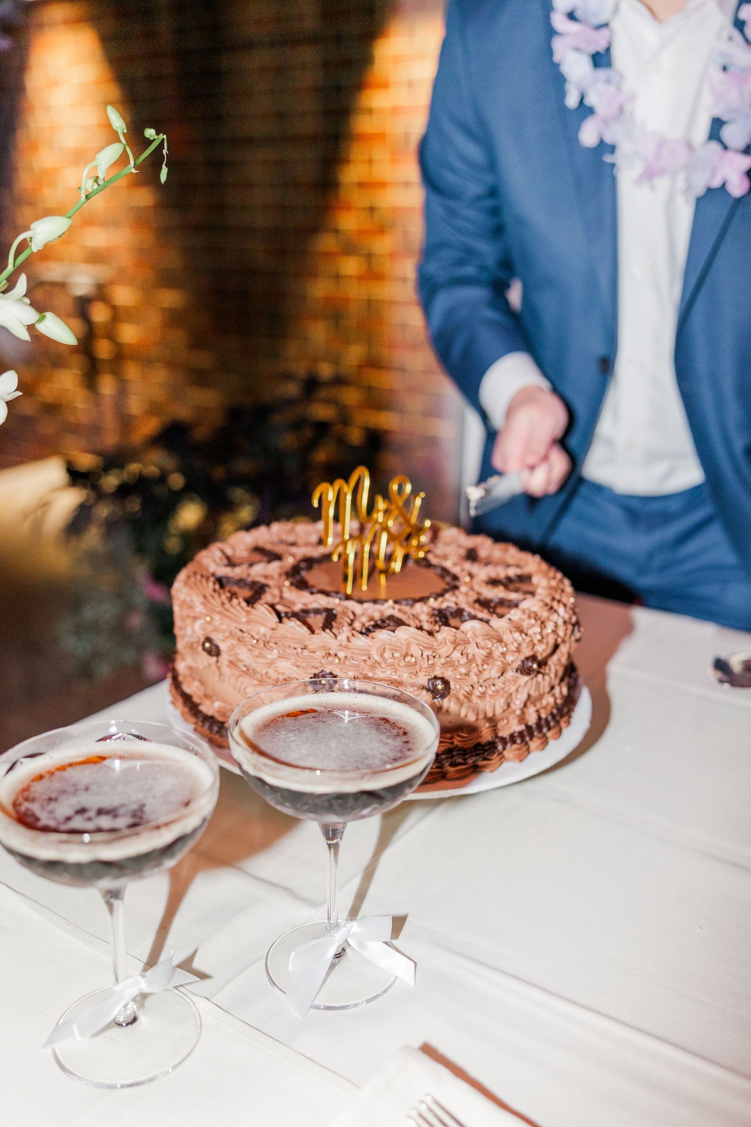 Chocolate frosted birthday cake with lit candles on white tablecloth next to empty dessert plates and wine glasses.