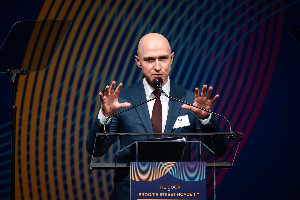 A speaker stands at a podium on stage with curved colorful light patterns in the background during an event.