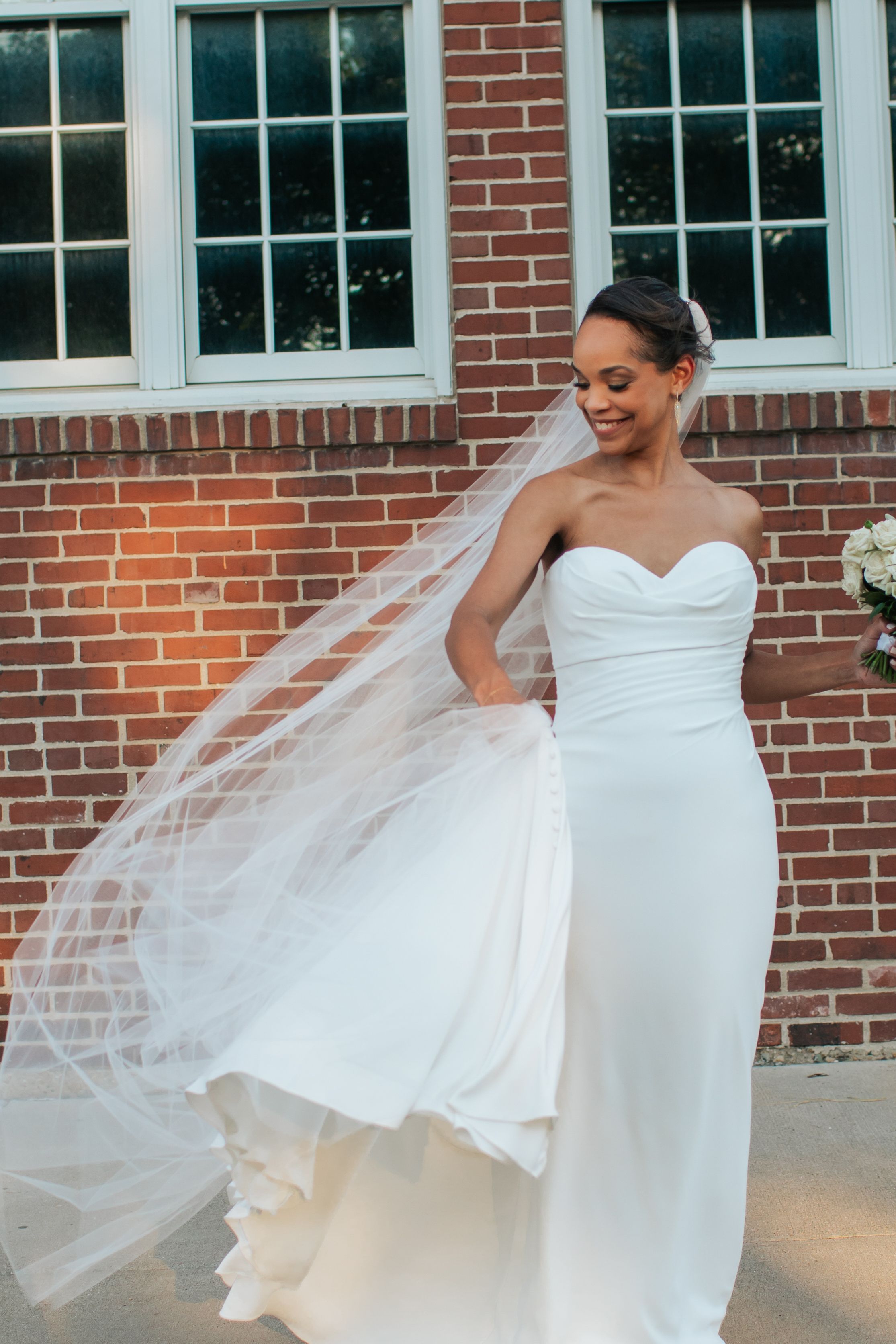 A bride in a white strapless wedding gown twirls gracefully, creating a flowing motion with her dress and veil against brick walls.