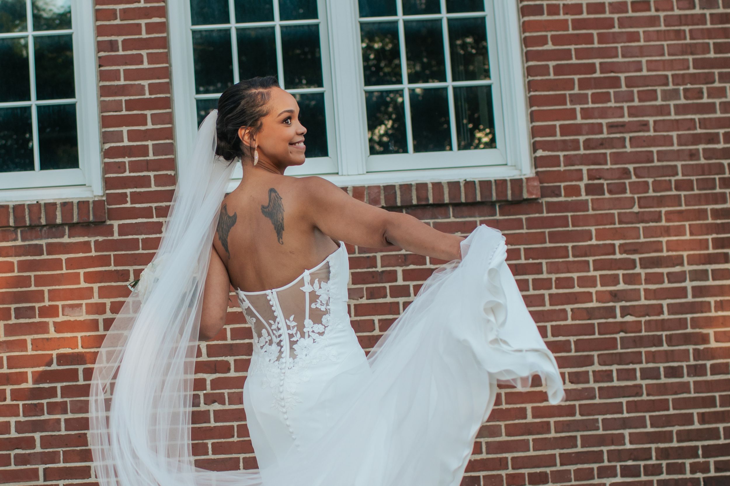 Bride in a flowing lace wedding gown poses playfully in front of a brick building with white windows.